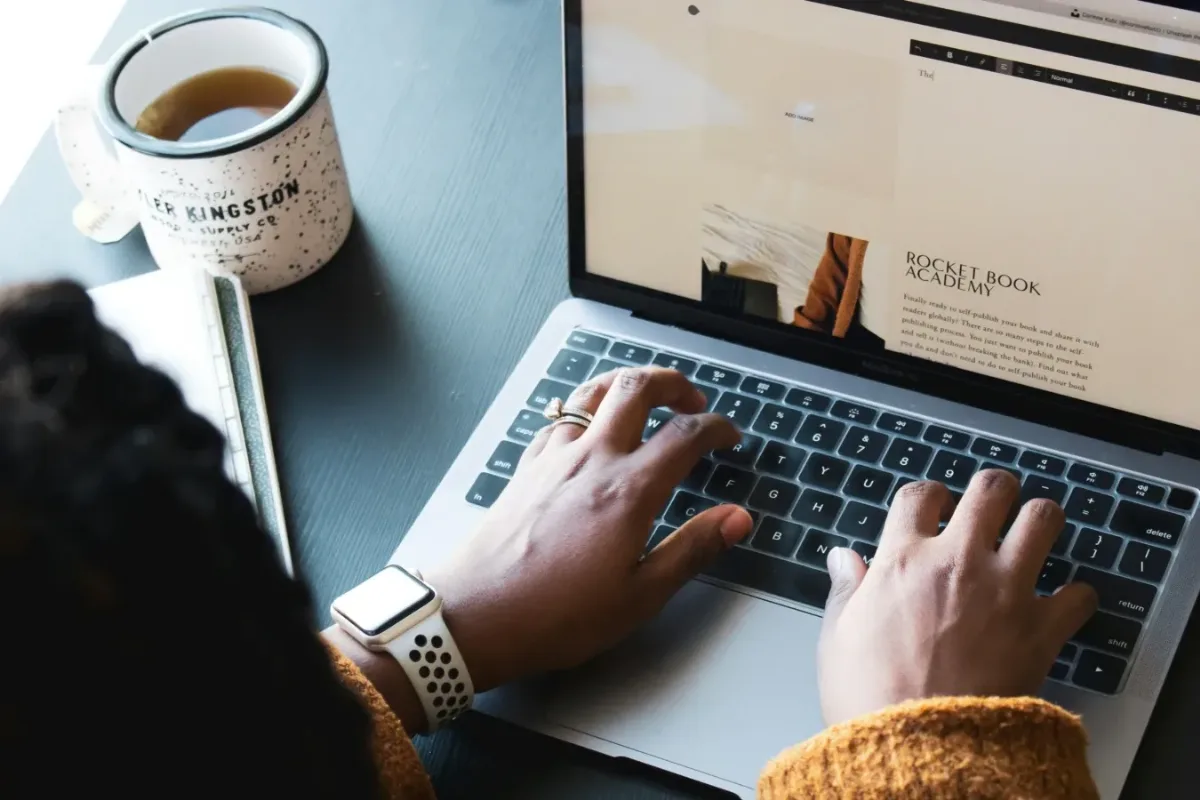 Person tippt auf Laptop-Tastatur neben Kaffeetasse mit Kingston-Aufdruck, Monitor zeigt Rocket Book Academy Website